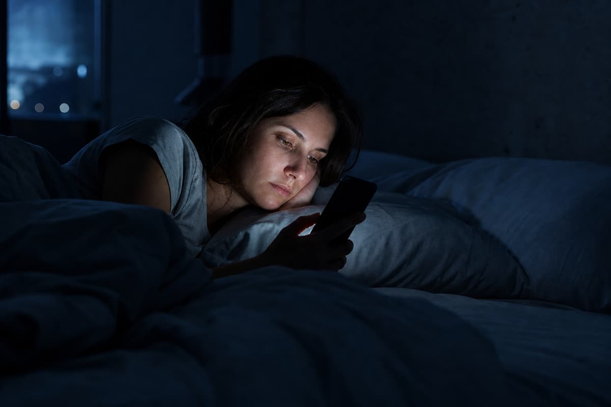 Tired person lying in bed at night, face illuminated by phone screen glow while doomscrolling